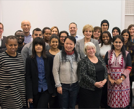 A diverse group of people gathered together in a room, posing for a group photo with smiles. They are dressed in casual and business attire.