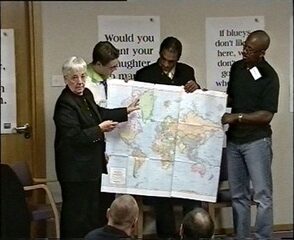 Jane Elliot wearing a black suit, holding a Peter's map, standing next to a group of students in a scene from the educational video series Perception is Everything.
